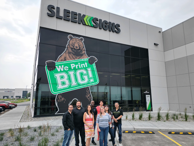 Sleek Signs employees stand in front of the office building with a sign that reads "We Print Big" held by a bear. 